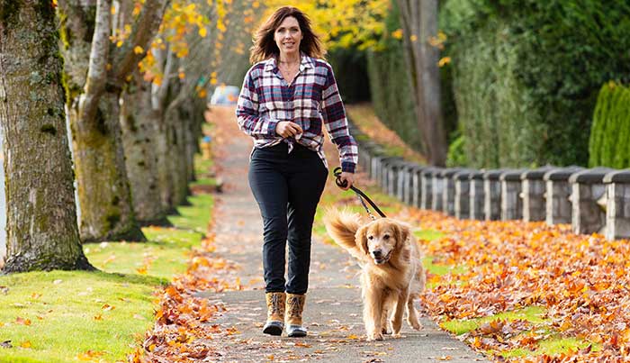 Woman walking a Golden Retriever dog on a tree-lined sidewalk covered in autumn leaves